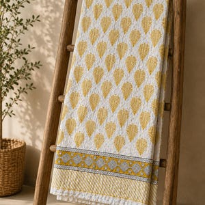 May include: A white quilted throw blanket featuring a repeating pattern of yellow leaves. The bottom edge displays a horizontal border with a grey and yellow geometric diamond design, finished with a thin yellow striped band at the very bottom hem.