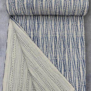 May include: A folded textile featuring a repeating dark blue leaf-like pattern on an off-white background. The fabric displays visible rows of straight running stitches in a contrasting dark thread, creating a textured, quilted appearance throughout.