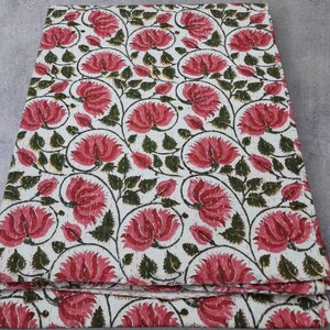 May include: Folded white fabric with a repeating pattern of large pink flowers, green leaves, and dark stems. The floral design is evenly spaced across the fabric's surface, creating a visually appealing textile.