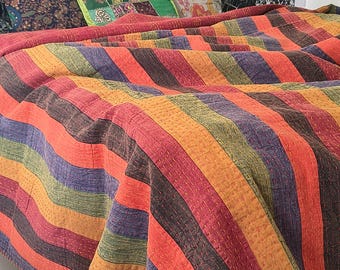 May include: A quilted bedspread featuring vertical stripes in shades of orange, yellow, purple, and green. The fabric displays visible running stitch details throughout the surface, creating a textured appearance across the entire length of the textile.
