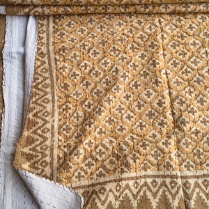 Op de afbeelding: Een gevouwen quilt met een herhalend bloemenpatroon in tinten bruin en beige. De quilt heeft een witte achterkant en is gestikt met bruin garen. De randen hebben een geometrische rand.