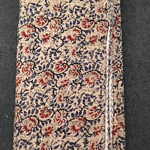 Op de afbeelding: Een gevouwen rechthoekig textiel met een crèmekleurige basis en een herhalend bloemenpatroon in tinten dieprood, marineblauw en zwart. De stof vertoont overal zichtbare lineaire stiksels, wat zorgt voor een gewatteerde textuur op het oppervlak.