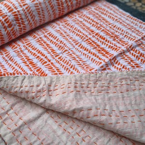 Puede incluir: Un edredón blanco y naranja estampado, parcialmente enrollado, con un diseño repetido de formas de gotas de color naranja. El edredón está cosido a mano con hilo naranja y los bordes son visibles.