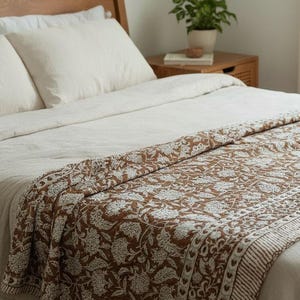 Op de afbeelding: Een bed met witte lakens en een bruin en wit bloemenpatroon quilt. De quilt heeft een herhalend bloemenontwerp en een gestreepte rand. Een houten hoofdbord en nachtkastje zijn zichtbaar.