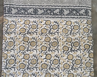May include: A folded, floral-patterned quilt. The quilt features a white background with a repeating pattern of yellow and black flowers and leaves. The top border has a striped design, and the middle border has a paisley design. The quilt is made of a soft material.