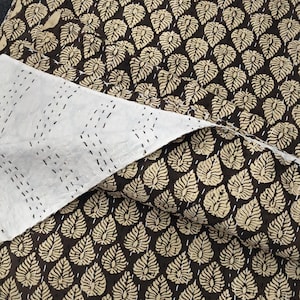 May include: A quilted textile featuring a dark brown background with a repeating pattern of light beige leaf shapes. The reverse side is solid white with visible rows of straight black running stitches that create a textured, quilted appearance.