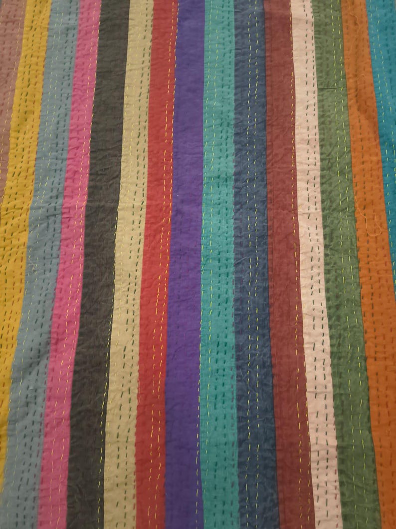 May include: A colorful textile with vertical stripes in various colors, including yellow, pink, red, purple, and teal. The fabric is stitched with yellow thread, creating a textured appearance. The overall design is vibrant and eye-catching.