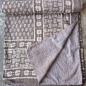 May include: A rolled-up, gray and white patterned quilt with a folded-over corner. The quilt features a repeating geometric design in white on a gray background. The reverse side is a solid gray color. The quilt is made of a soft, quilted fabric.