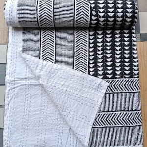 May include: A rolled-up, black and white patterned quilt with a geometric design. The quilt is partially unrolled, revealing a white interior with stitched details. The pattern includes vertical lines, chevrons, and triangles.