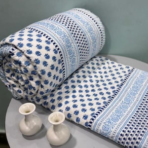 May include: Rolled-up white and blue patterned quilt with a floral design. The quilt is partially unrolled, revealing more of the pattern. Two small white vases are in the foreground. The quilt's design features a repeating floral pattern.