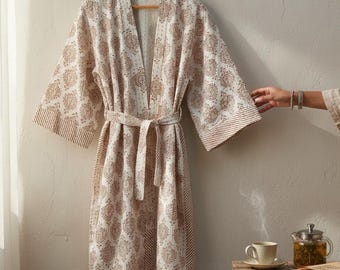 May include: A white and brown patterned robe hangs on a wooden hanger. The robe has a matching belt and a repeating floral design. The sleeves and edges are trimmed with a striped pattern. A hand with bracelets is visible.