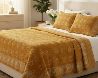 May include: Mustard yellow quilt and two matching pillow shams with a floral pattern. The bedding set features a detailed border design and is placed on a bed with a neutral headboard. The room has a bright, airy feel.