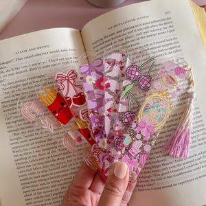 Puede incluir: Una colección de marcadores de acrílico transparente con varios diseños, incluyendo cerezas, papas fritas y patrones florales. Cada marcador tiene una forma y esquema de color únicos, con acentos rosas y morados. Los marcadores se muestran en un libro abierto.