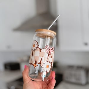 Puede incluir: Vaso de vidrio transparente con forma de lata con tapa de madera y pajita de metal. El vaso presenta un diseño con la letra "M", margaritas, un lazo rosa y un estampado de cuadros en tonos marrones y beige.