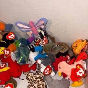 May include: A selection of colourful plush toys, including a butterfly, badger, toucan, and rooster. Each toy has a tag attached. The toys are in a box, with a white background.