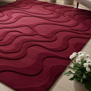 Puede incluir: Una alfombra rectangular grande de color burdeos con un patr&oacute;n ondulado en relieve. El dise&ntilde;o presenta una serie de formas interconectadas en forma de ola en la superficie. La alfombra est&aacute; hecha de un material suave y texturizado y est&aacute; colocada sobre un suelo de madera clara.