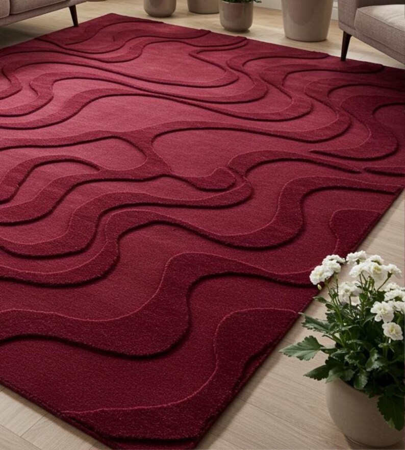 Puede incluir: Una alfombra rectangular grande de color burdeos con un patr&oacute;n ondulado en relieve. El dise&ntilde;o presenta una serie de formas interconectadas en forma de ola en la superficie. La alfombra est&aacute; hecha de un material suave y texturizado y est&aacute; colocada sobre un suelo de madera clara.