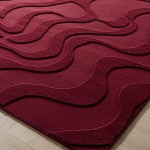 Puede incluir: Una alfombra burdeos con un patr&oacute;n ondulado en relieve. El dise&ntilde;o de la alfombra presenta l&iacute;neas fluidas en diferentes tonos de burdeos, creando un efecto texturizado. La alfombra est&aacute; colocada sobre un suelo de madera clara.