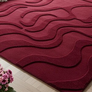 Puede incluir: Alfombra burdeos con un dise&ntilde;o ondulado. La alfombra tiene una superficie texturizada con diferentes tonos de rojo, creando un efecto tridimensional. Se encuentra sobre un suelo de madera clara.