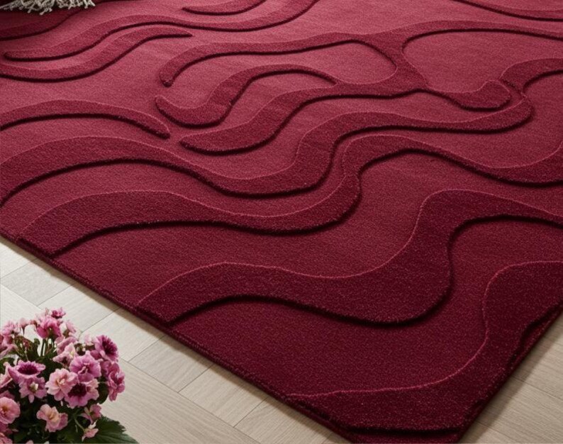 Puede incluir: Alfombra burdeos con un dise&ntilde;o ondulado. La alfombra tiene una superficie texturizada con diferentes tonos de rojo, creando un efecto tridimensional. Se encuentra sobre un suelo de madera clara.