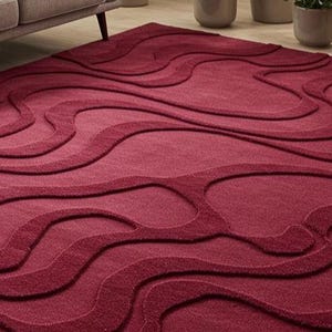 Puede incluir: Una alfombra de color burdeos con un dise&ntilde;o ondulado en relieve. El dise&ntilde;o de la alfombra presenta una serie de formas interconectadas en forma de onda en diferentes tonos de rojo. La alfombra est&aacute; colocada sobre un suelo de madera, con un sof&aacute; y una manta en el fondo.