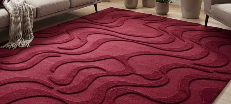 Puede incluir: Una alfombra de color burdeos con un dise&ntilde;o ondulado en relieve. El dise&ntilde;o de la alfombra presenta una serie de formas interconectadas en forma de onda en diferentes tonos de rojo. La alfombra est&aacute; colocada sobre un suelo de madera, con un sof&aacute; y una manta en el fondo.