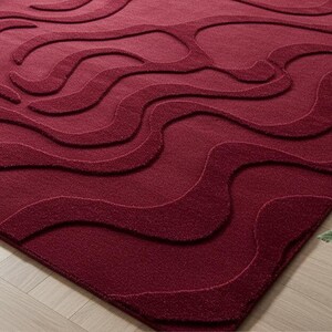 Puede incluir: Una alfombra burdeos con un dise&ntilde;o de ondas. La alfombra tiene una superficie texturizada y un color rojo intenso. El dise&ntilde;o presenta l&iacute;neas onduladas en relieve que crean una sensaci&oacute;n de movimiento y profundidad. La alfombra es de forma rectangular.