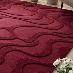 Puede incluir: Una alfombra de color burdeos con un dise&ntilde;o en relieve y fluido. El dise&ntilde;o de la alfombra presenta una serie de formas onduladas interconectadas, creando una superficie texturizada. La alfombra est&aacute; hecha de un material suave.