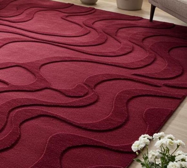Puede incluir: Una alfombra de color burdeos con un dise&ntilde;o en relieve y fluido. El dise&ntilde;o de la alfombra presenta una serie de formas onduladas interconectadas, creando una superficie texturizada. La alfombra est&aacute; hecha de un material suave.
