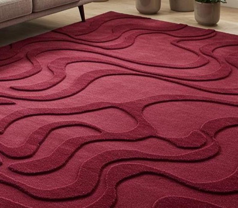 Puede incluir: Una alfombra burdeos con un patr&oacute;n en relieve y fluido. El dise&ntilde;o de la alfombra presenta una serie de formas onduladas interconectadas, creando una superficie texturizada. La alfombra est&aacute; hecha de un material suave y es adecuada para una sala de estar o un dormitorio.