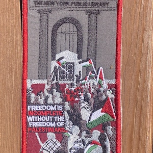 May include: A rectangular embroidered patch with a red border featuring a gray building labeled THE NEW YORK PUBLIC LIBRARY. Below, a crowd holds Palestinian flags and a sign reading CEASE FIRE. Text reads FREEDOM IS INCOMPLETE WITHOUT THE FREEDOM OF PALESTINIANS.