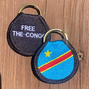 May include: Two embroidered keychains with gold-colored rings. One is black with the text "FREE THE CONGO" in white. The other features the flag of the Democratic Republic of the Congo: a blue field with a yellow star and a red stripe.