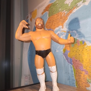 May include: A plastic action figure of a professional wrestler with brown hair and a beard, wearing black trunks and white lace-up wrestling boots with knee pads. The figure is posed with one arm flexed and the other arm extended to the side.
