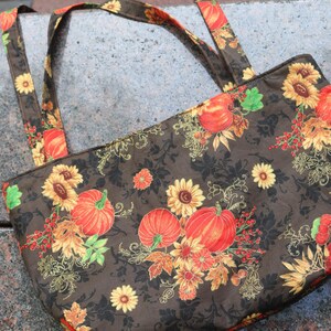 Könnte beinhalten: Eine braune Tragetasche mit einem herbstlichen Muster. Der Stoff zeigt Kürbisse, Sonnenblumen und Herbstblätter in Orange-, Gelb- und Rottönen auf dunklem Hintergrund. Die Tasche hat passende Griffe.