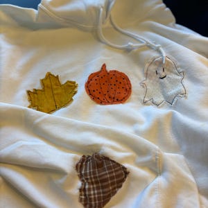 Könnte beinhalten: Weißer Kapuzenpullover mit Applikationen zum Thema Herbst. Mit einem gelben Blatt, einem orangefarbenen Kürbis, einem weißen Geist und einem braunen karierten Herz. Der Kapuzenpullover hat eine Kapuze mit Kordelzug und eine Vordertasche.