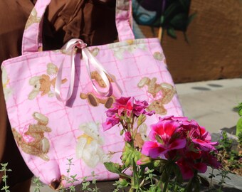 May include: A pink tote bag with a repeating pattern of teddy bears and bunnies. The bag has a quilted design and pink ribbon ties. The handles are also pink. The bag is next to pink and red flowers.