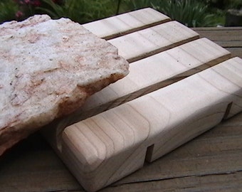 May include: A light-colored wooden soap dish with a slatted design, paired with a rough-hewn, white and tan stone. The soap dish has a rectangular shape with four raised slats and cut-out slots for drainage. The stone has a natural, textured appearance.