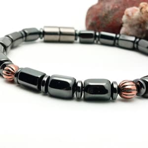 May include: A dark gray magnetic bracelet with rectangular and cylindrical beads. The bracelet features copper-colored, spherical accents. The bracelet is displayed on a white surface with a blurred background.