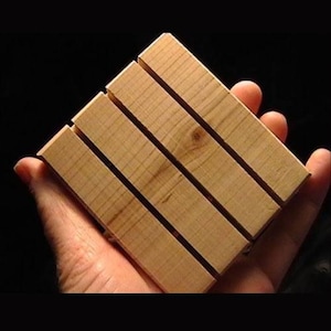 May include: A square wooden soap dish made of light-colored wood. The dish is constructed from four parallel wooden slats, with narrow gaps between them. The wood grain is visible, and the dish is held in a hand against a dark background.