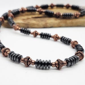May include: A necklace featuring black, cylindrical beads interspersed with copper-colored, decorative elements. The necklace is displayed on a white surface, with a wooden element in the background. The design is symmetrical and the beads are evenly spaced.