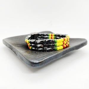 May include: A three-strand bracelet with black, faceted beads and colorful accents. The bracelet features yellow, red, and green beads, resting on a dark gray square plate. The bracelet is a fashion accessory.
