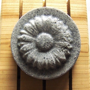 May include: A round, gray soap bar with a raised, flower-like design. The soap has a textured surface and a slightly darker gray center.