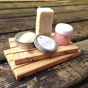May include: A set of three natural bath products on a wooden soap dish. The set includes a bar of soap, a small jar of lip balm, and a tin of body butter. The soap is white and has a natural scent. The lip balm is pink and has a sweet scent. The body butter is white and has a creamy texture.