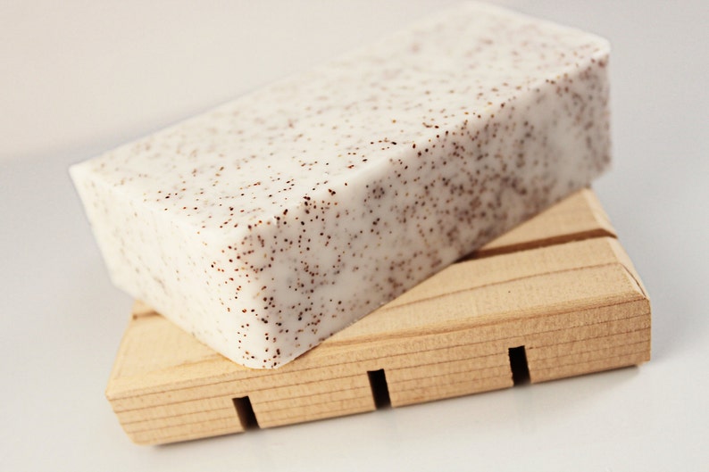 K&ouml;nnte beinhalten: Ein rechteckiger Seifenblock mit gesprenkeltem Aussehen liegt auf einer Seifenschale aus Holz. Die Seife ist cremefarben mit kleinen braunen Punkten. Die Holzschale hat eine nat&uuml;rliche Oberfl&auml;che und ein geschlitztes Design. Geeignet f&uuml;r Kosmetik- und K&ouml;rperpflegeprodukte.