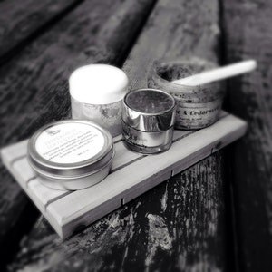 May include: A grayscale image of a wooden tray holding four cosmetic products. The products include a small round tin labeled "Peppermint Tooth Cream", a jar with a white lid, a silver jar labeled "City Unique", and a jar with a wooden spoon.