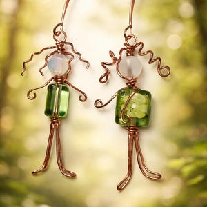 May include: Handmade copper wire earrings featuring whimsical figures. Each earring has a clear or opalescent bead head, a green glass body, and wire arms and legs. The earrings are designed to resemble small people.