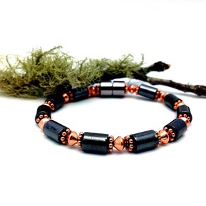 May include: A black and copper-colored bracelet with cylindrical and spherical beads. The bracelet features a magnetic clasp and is displayed on a white background with a sprig of moss and a twig.
