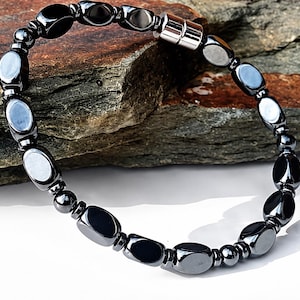 May include: A bracelet composed of polished, dark gray, oval-shaped hematite beads alternating with smaller, round, metallic beads. The piece features a cylindrical silver-toned magnetic clasp that connects the two ends of the beaded strand.