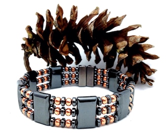 May include: A magnetic bracelet with a three-row design. The bracelet features rectangular and round beads in black and copper colors. A large brown pine cone is in the background.