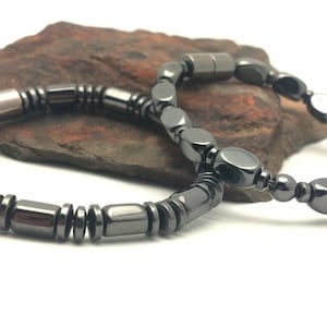 May include: Two black hematite bracelets, one with oval beads and one with rectangular beads, on a brown rock.